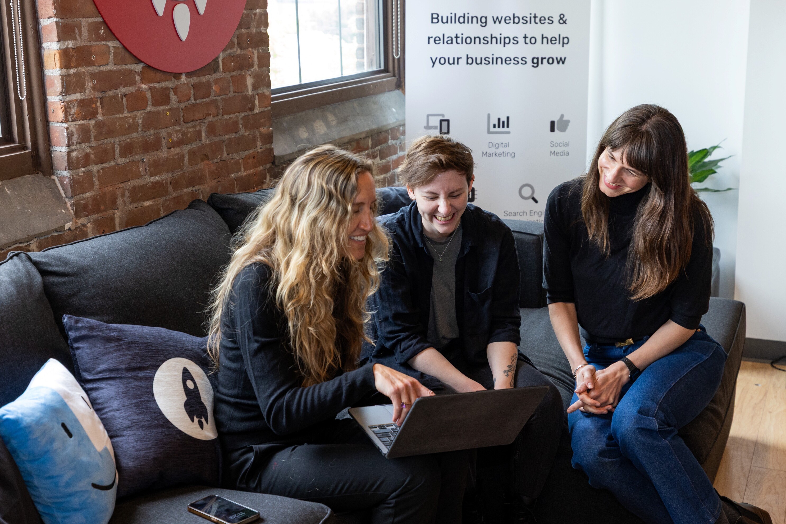 Three people collaborate on a laptop while seated on a sofa in a bright office setting.