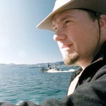 Man in a hat gazing out at water with a boat in the background.