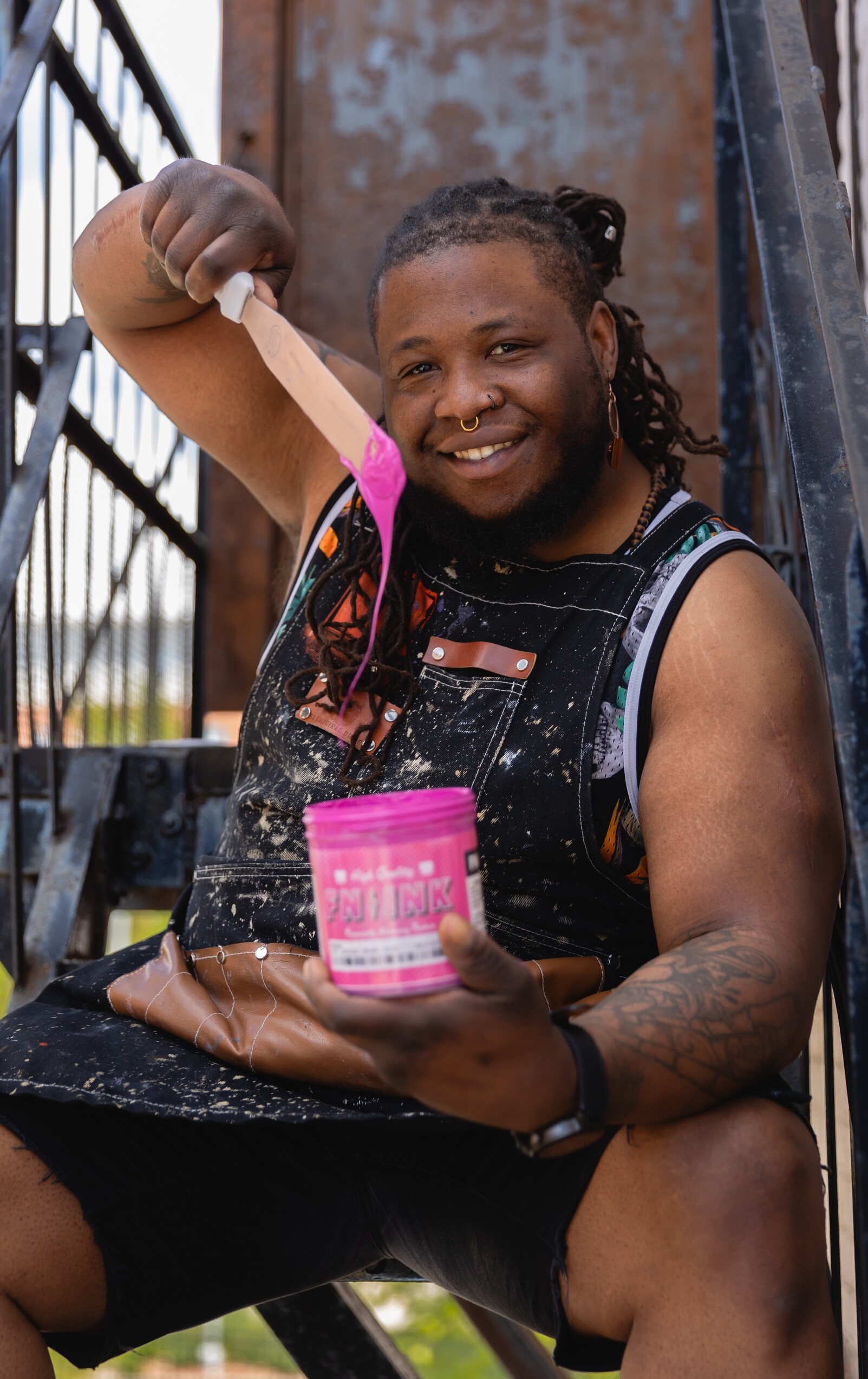A smiling person holds a paint scraper and a pink paint can while sitting on stairs.