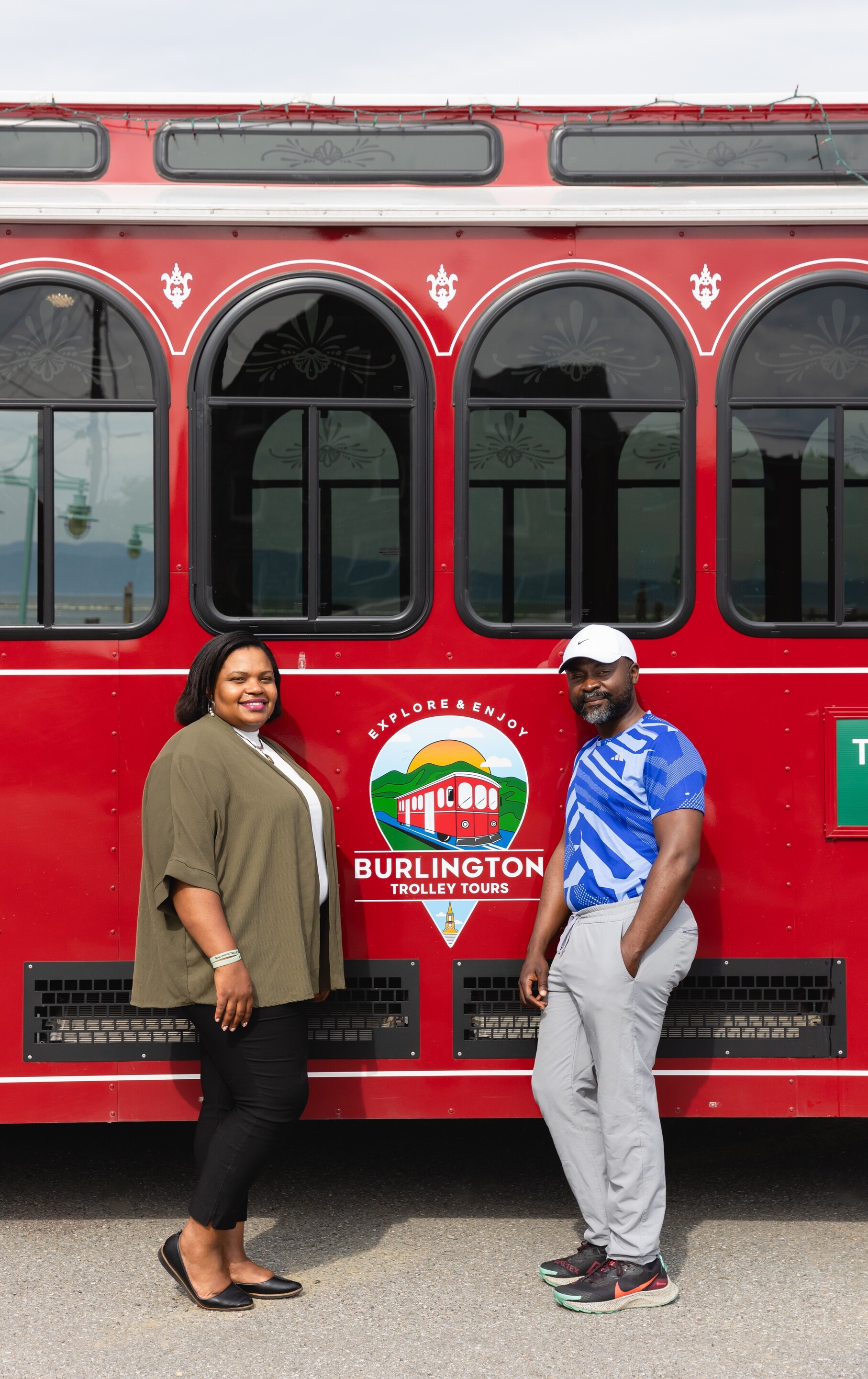 Two individuals pose next to a red trolley, promoting Burlington Trolley Tours.