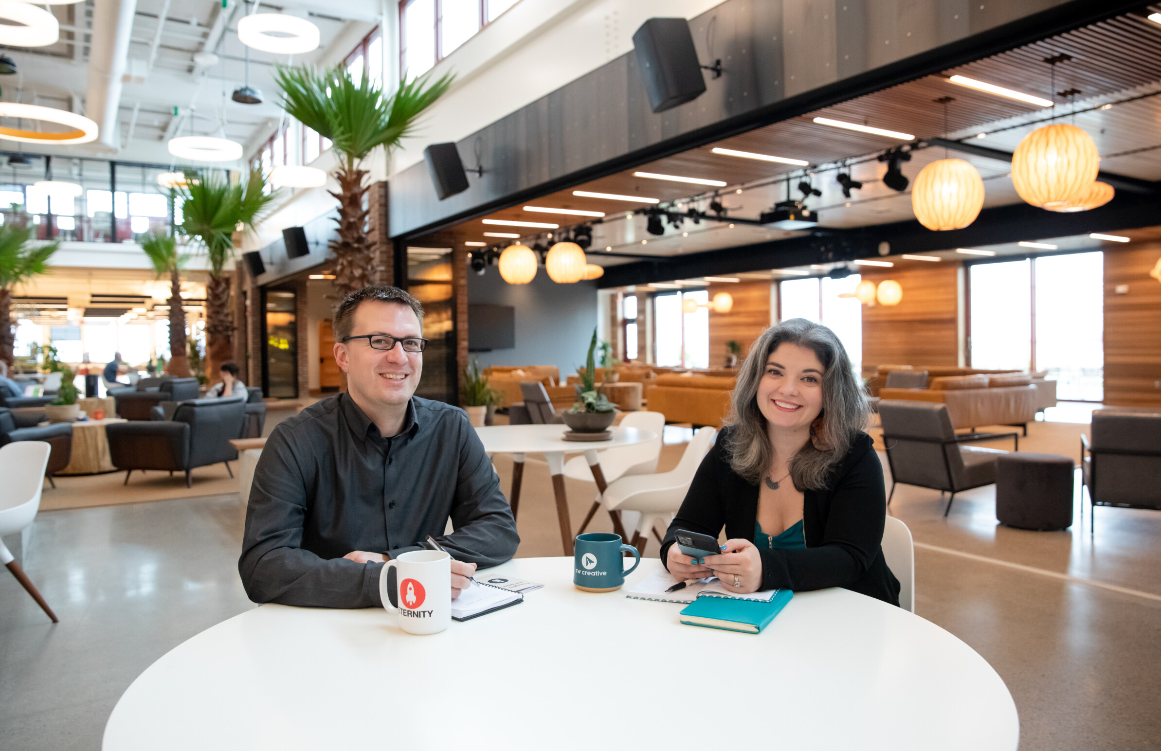 Two smiling professionals sit at a table in a modern workspace with greenery around.
