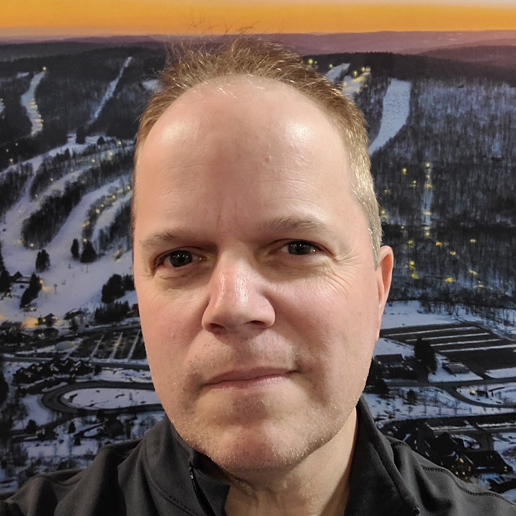 A man with short hair poses in front of a snowy landscape backdrop at sunset.