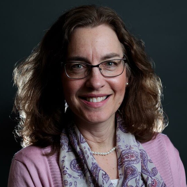 Woman with curly hair, glasses, and a scarf, smiling against a dark background.