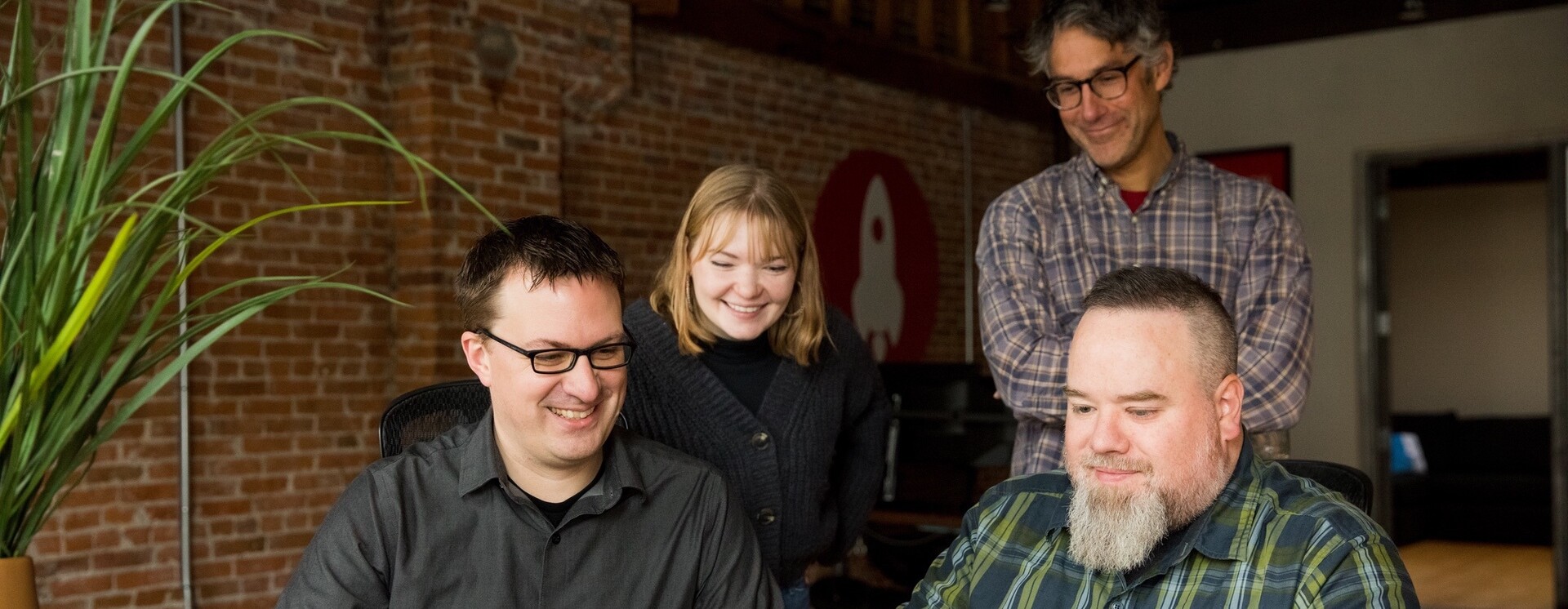 Four people are gathered around a computer, smiling and engaged in discussion.
