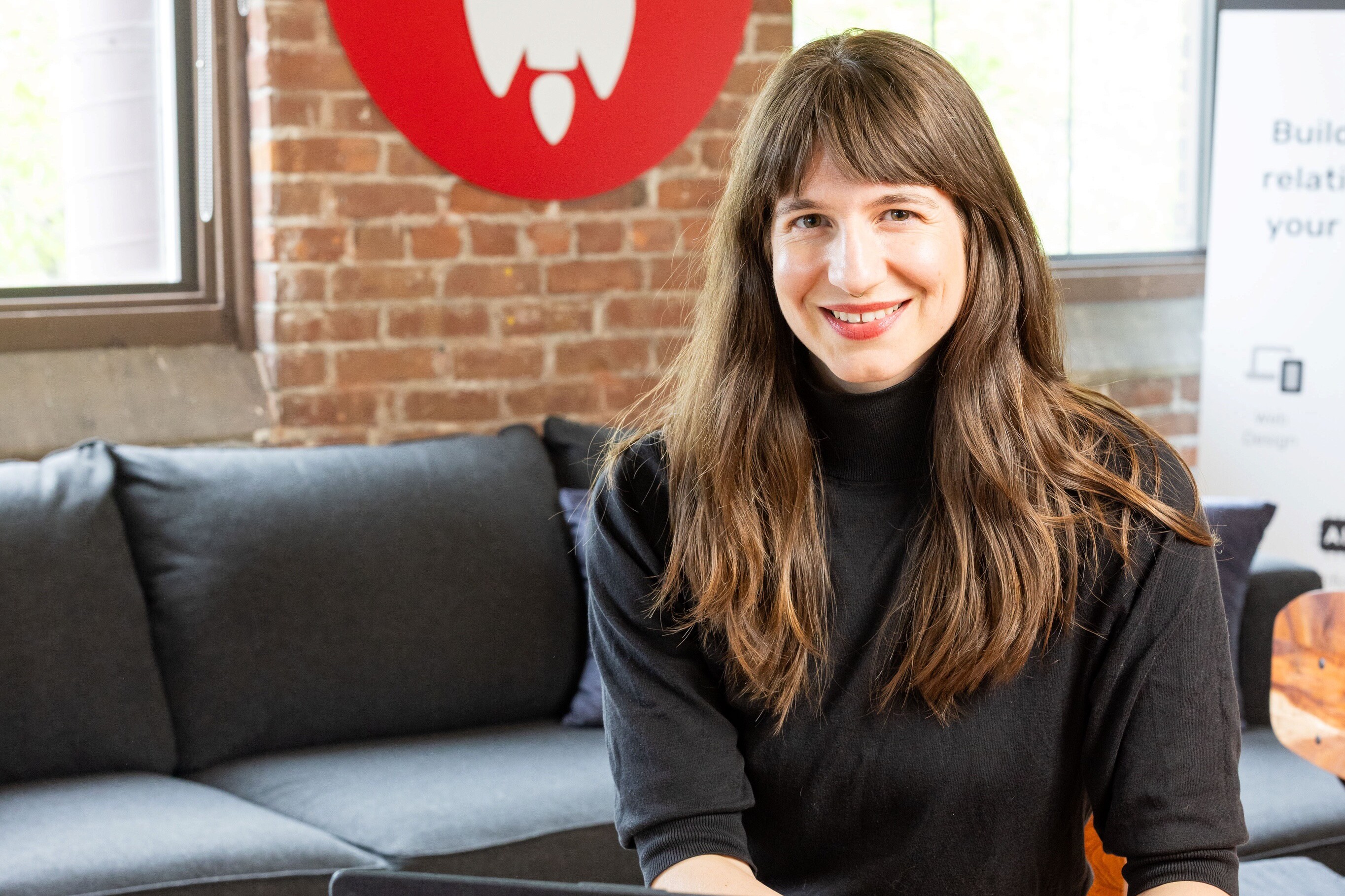 Smiling woman in a black shirt sits on a couch with a brick wall backdrop.