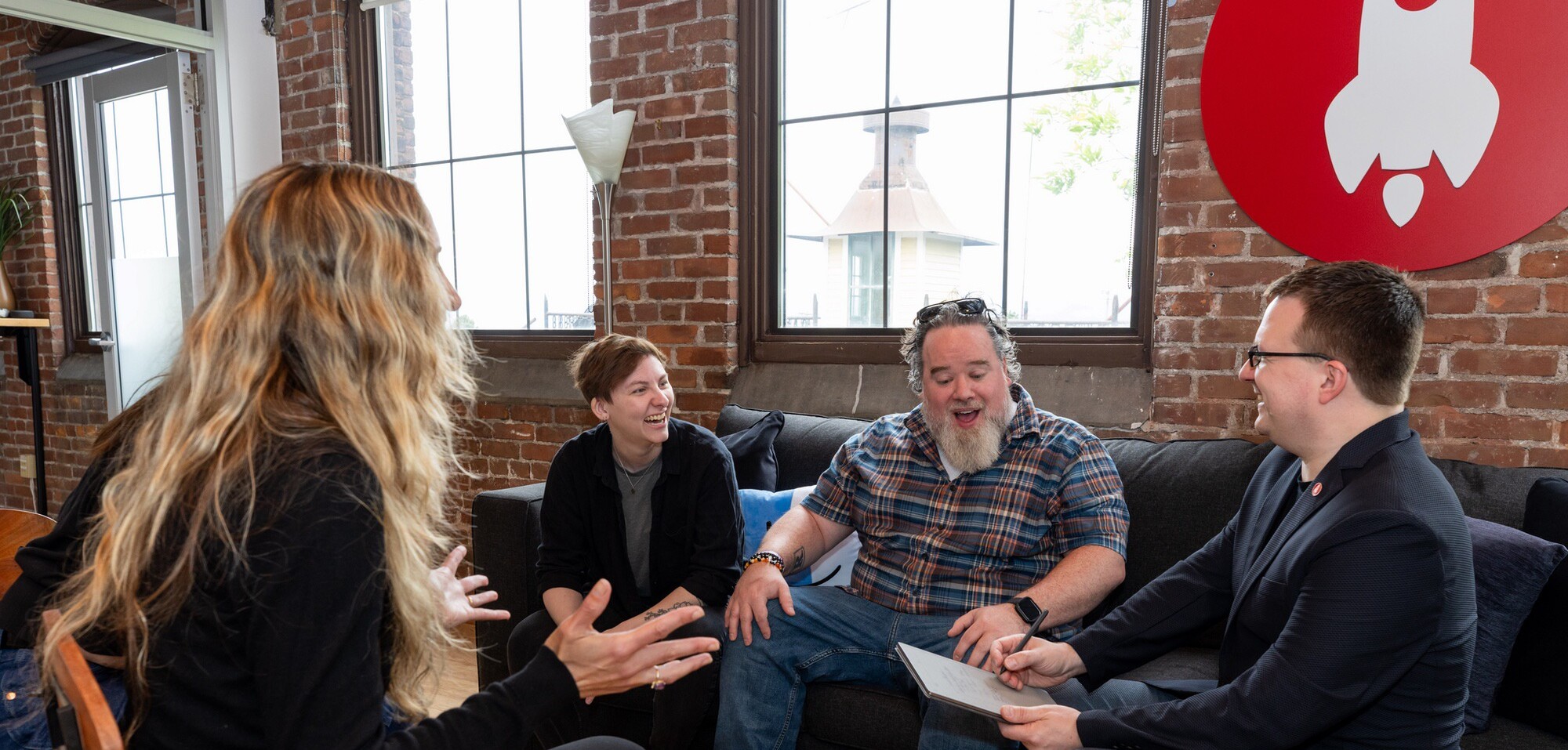 Four people engage in a lively discussion in a cozy, brick-walled office setting.