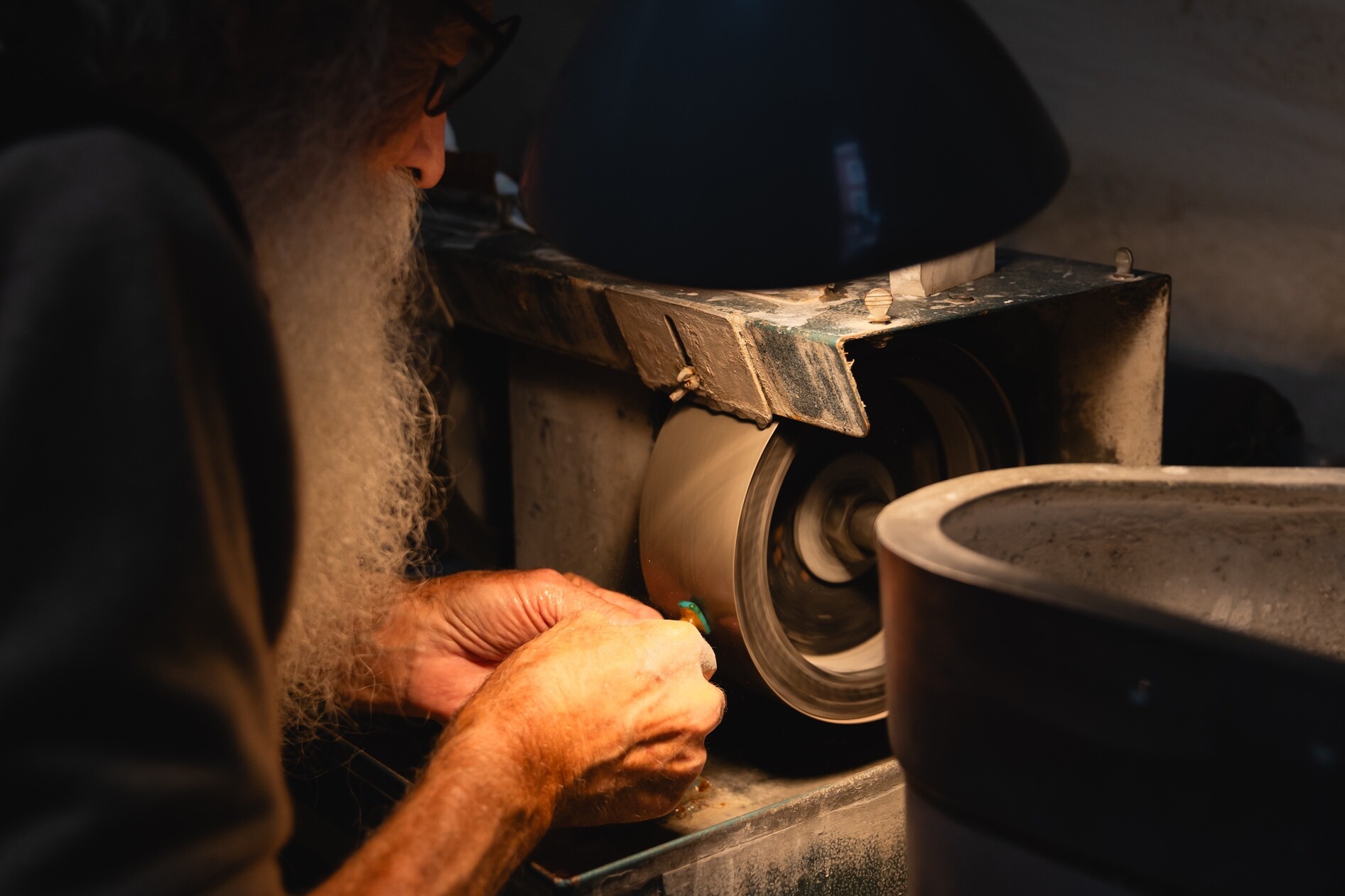 A person with a beard sharpens a tool at a workbench under a focused lamp.