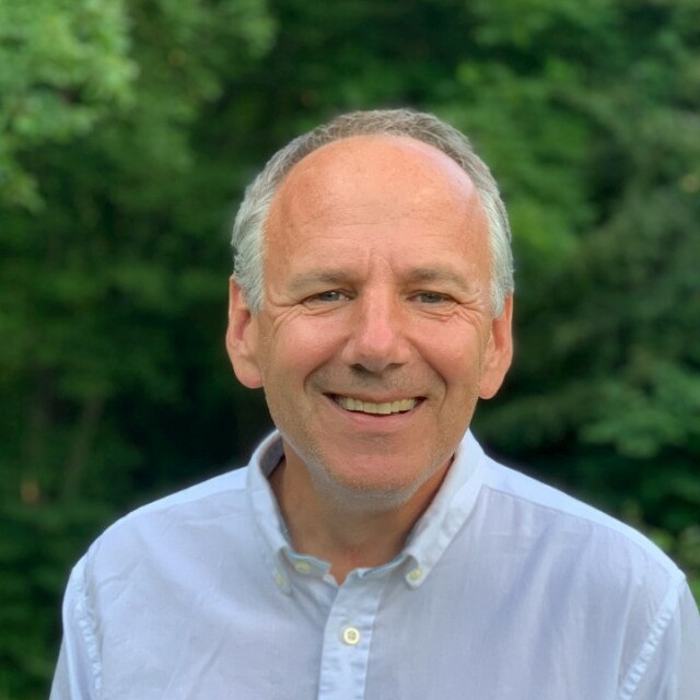 Smiling man in a light shirt stands against a green, natural backdrop.