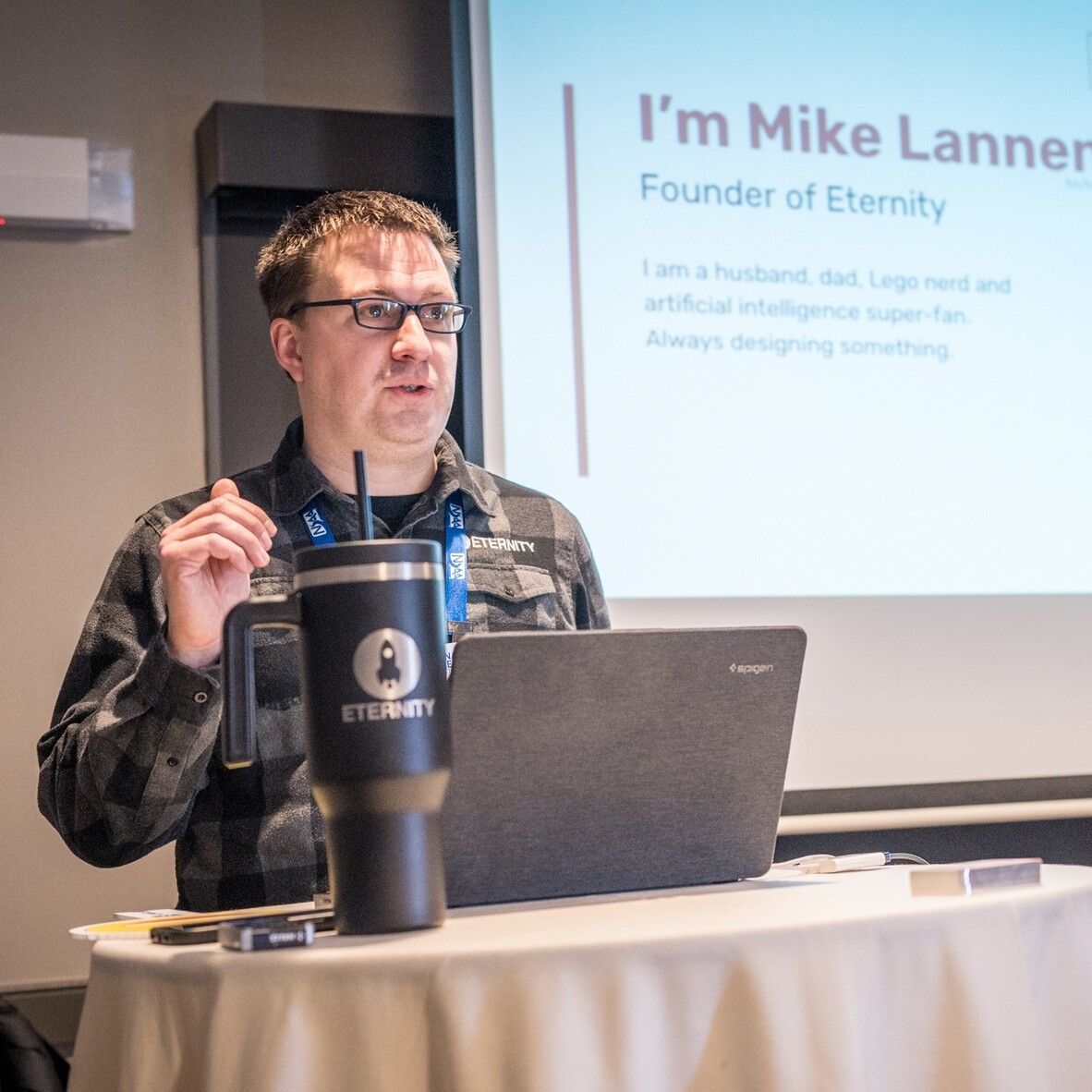 Speaker Mike Lanner presents about his work at a conference, with a coffee tumbler beside him.