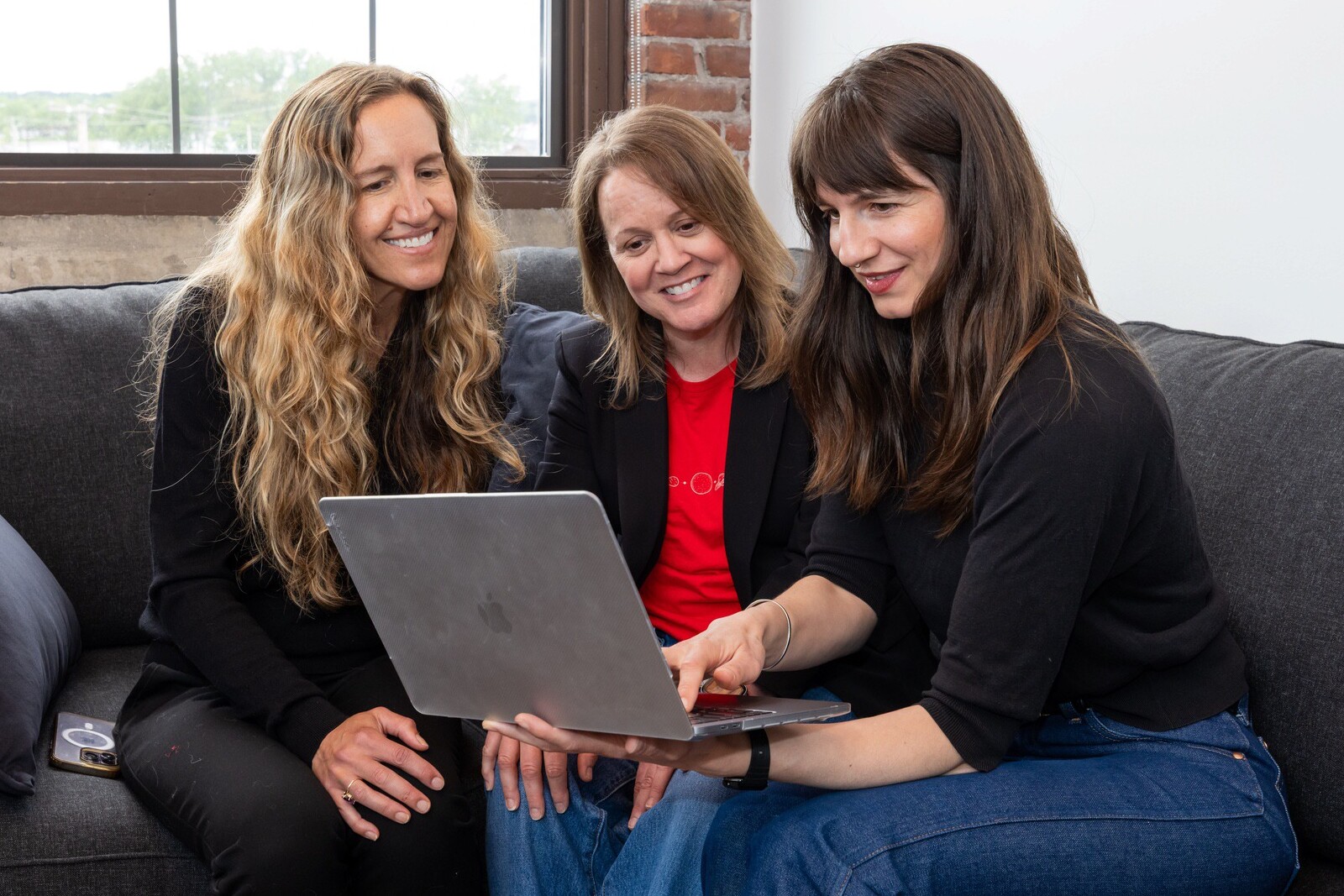 Three women sit together on a couch, looking at a laptop and smiling.