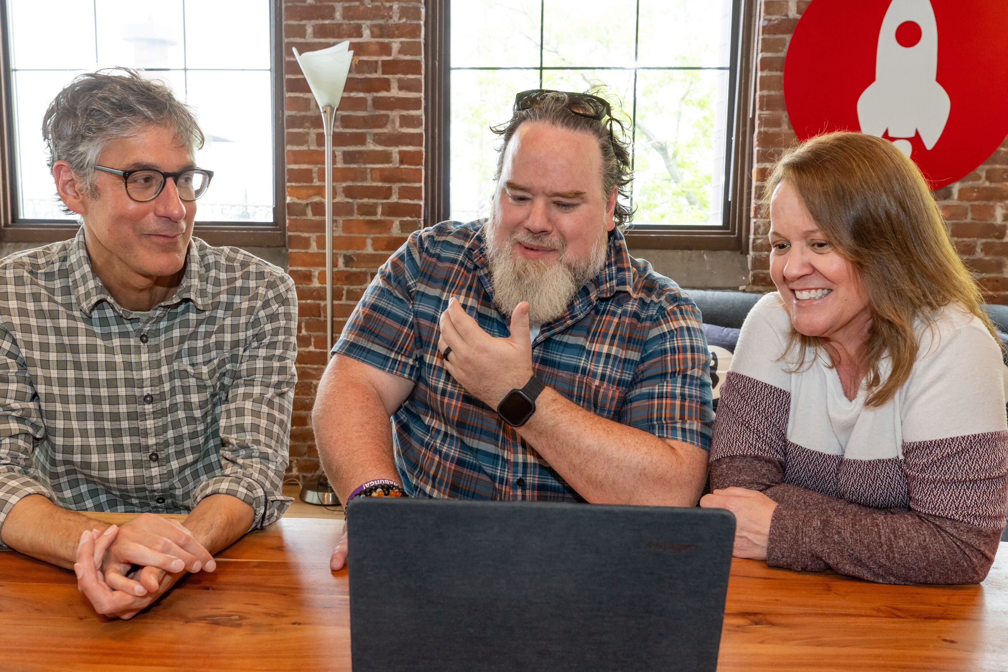 Three people sitting at a table, engaged and smiling while looking at a laptop.