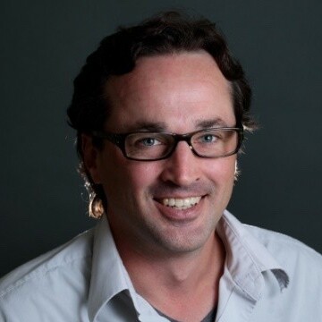 Man smiling, wearing glasses and a white shirt, against a dark background.