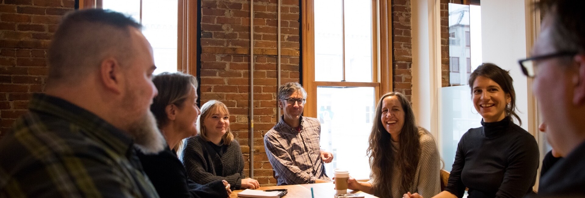 A group of eight people smiling and chatting around a table in a bright, brick-walled office.