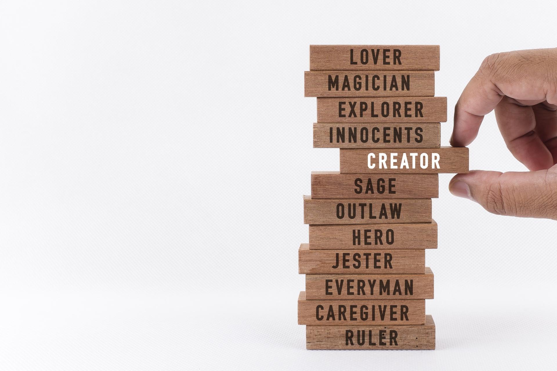wooden blocks stacked on top of each other with the words lover, magician, explorer, innocents, creator, sage, outlaw, hero, jester, everyman, caregiver, and ruler written individually on each. The creator block is being pulled out from the middle by a person's thumb and forefinger