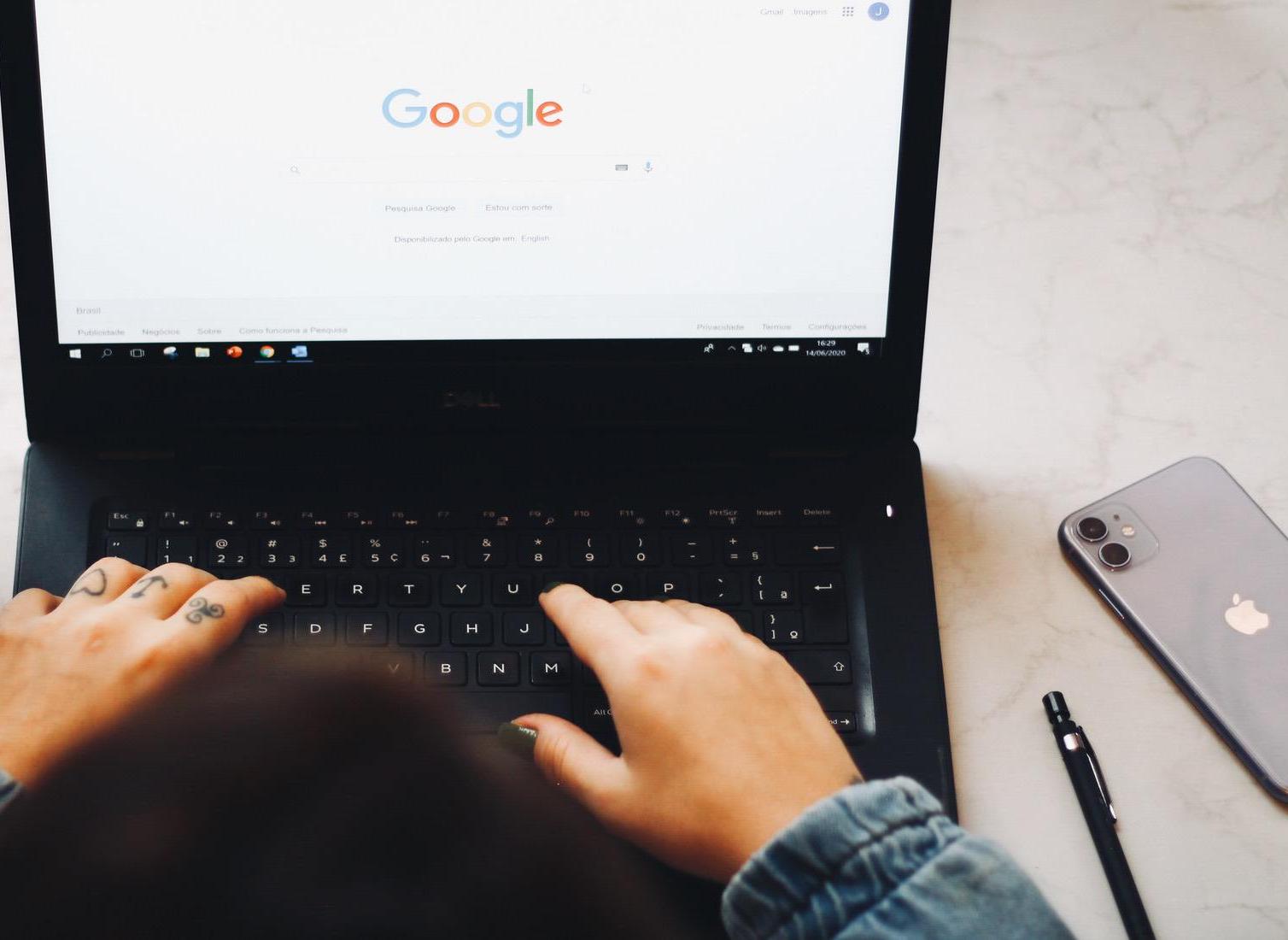 Woman typing into a google search on her laptop sitting at her desk with flowers to her left and her phone and a pen to her right
