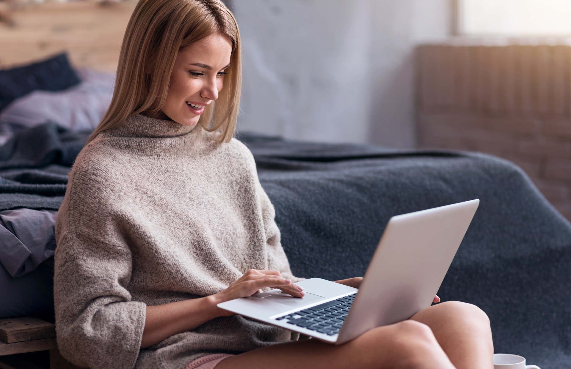 young woman in a sweater sitting at the foot of a bed smiling and looking at a laptop computer