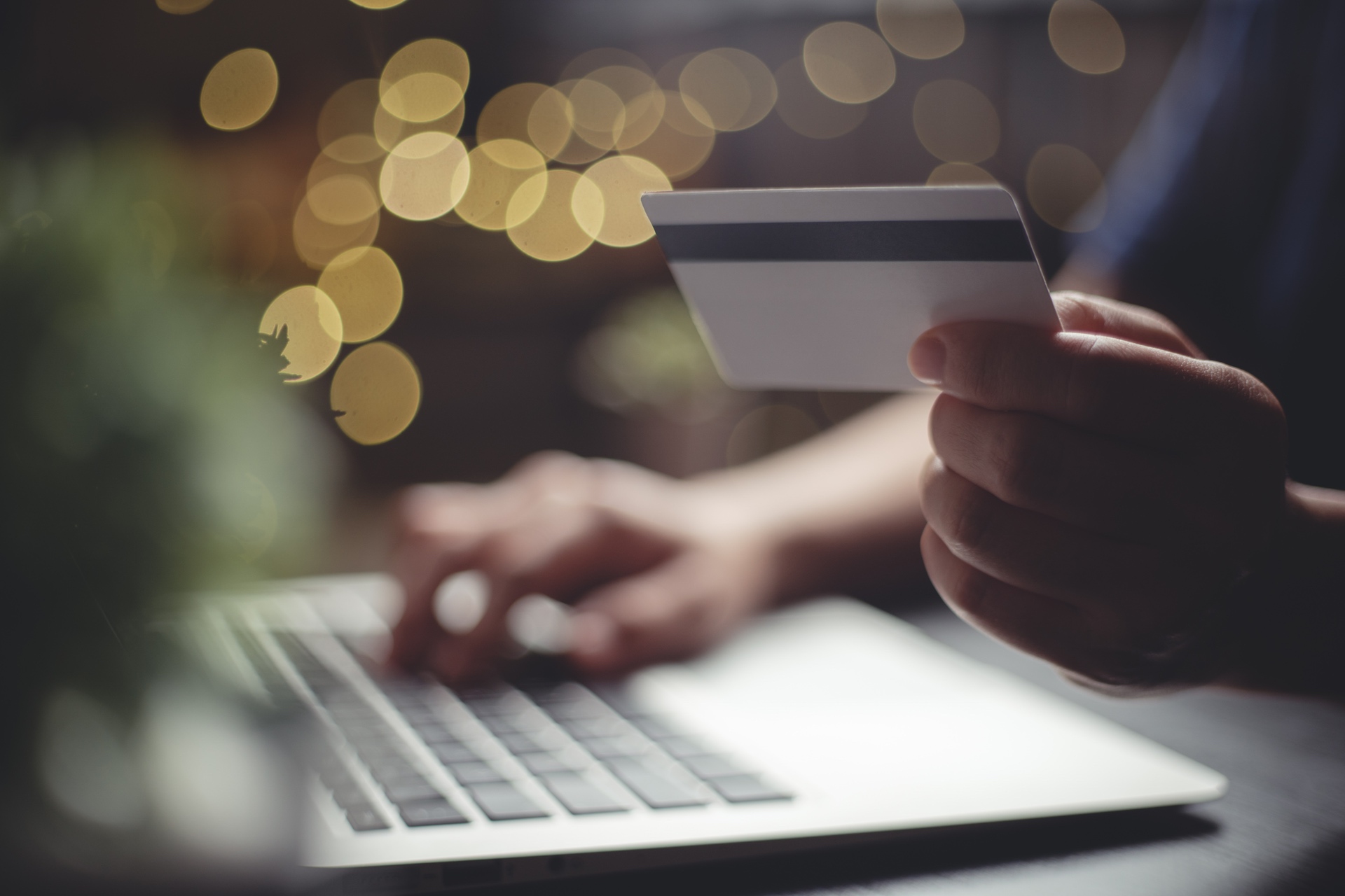 close up of a hand holding a credit card while the other hand types on a laptop at night with blurred lights in the background