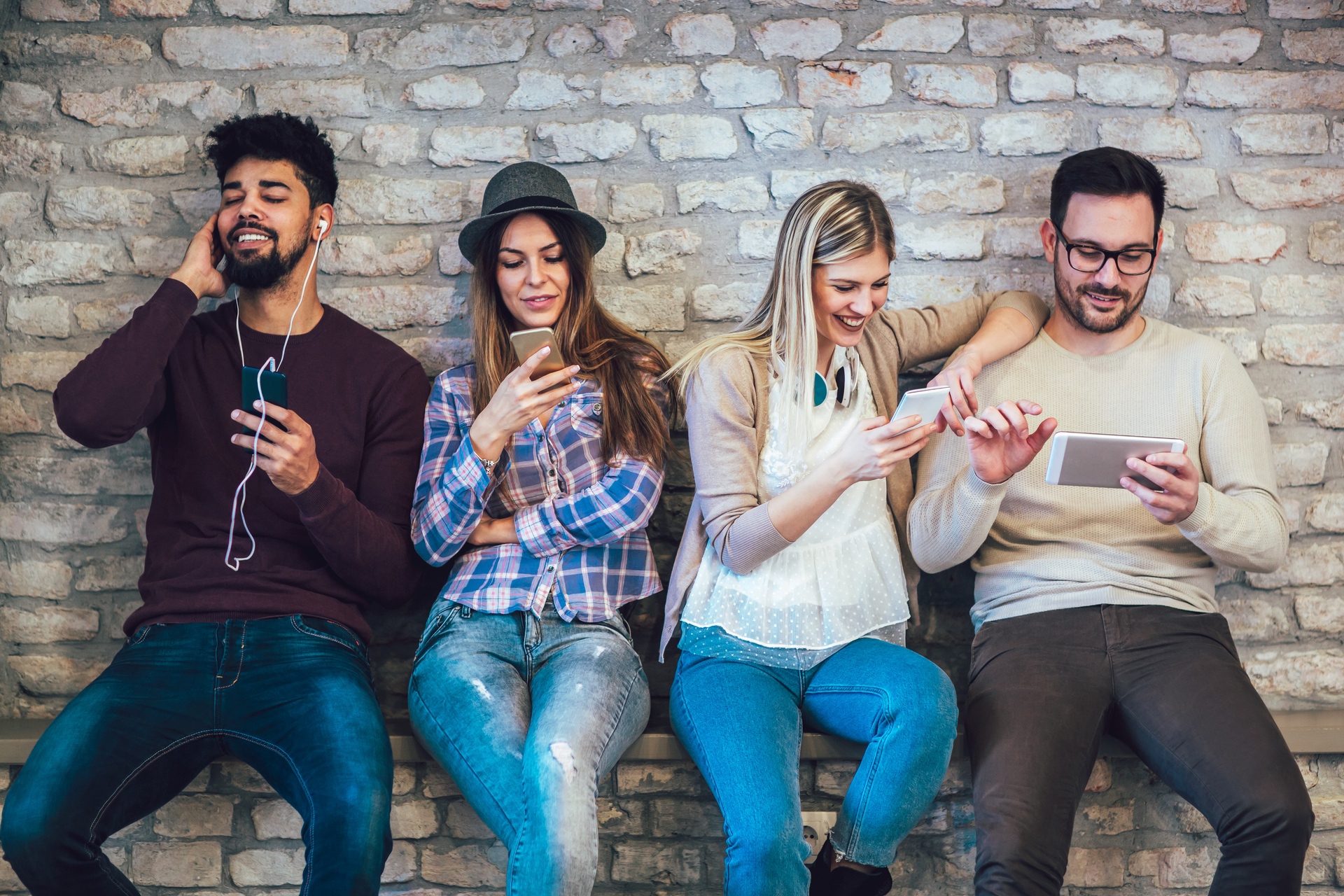 four young adults sitting on a bench against a wall, each using a cellphone